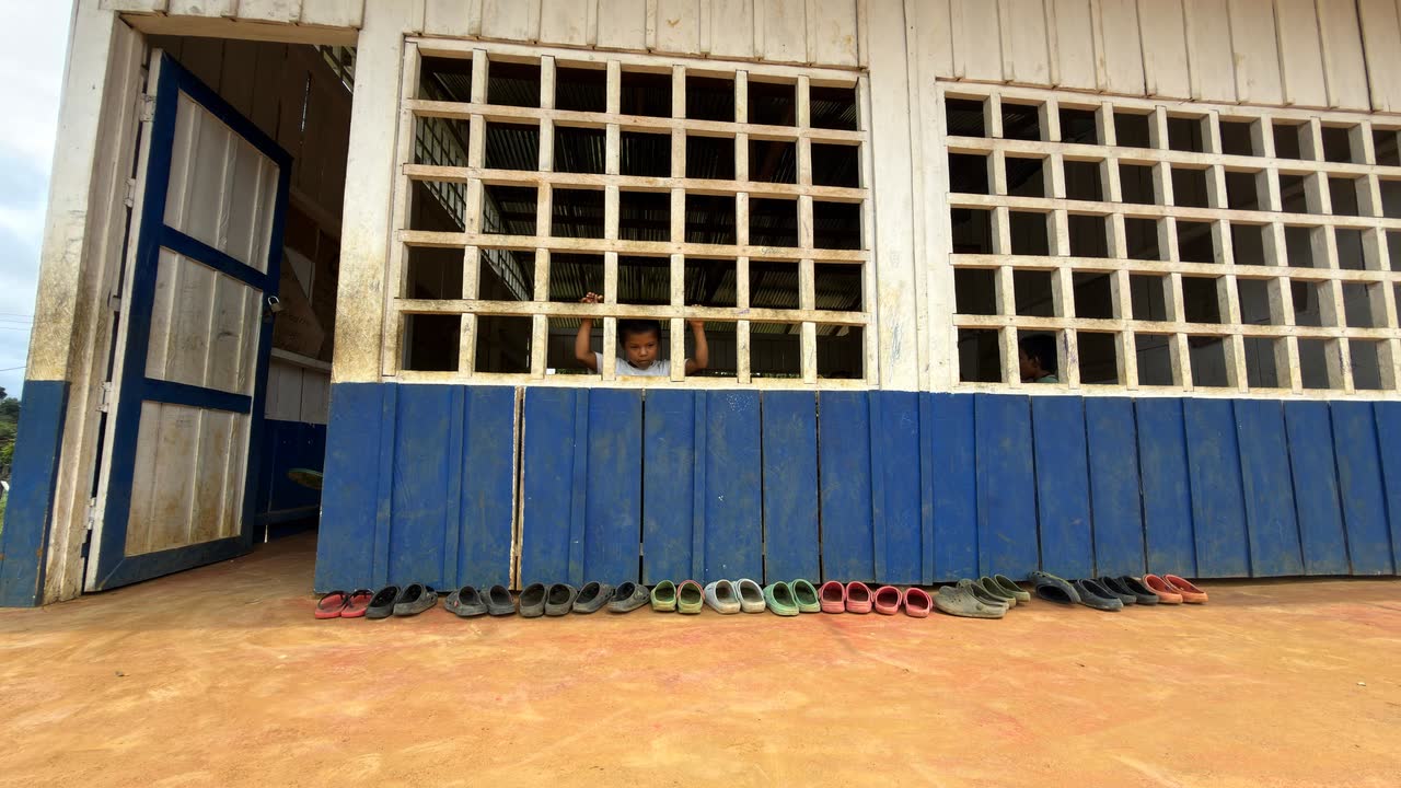 Zapatos alineados a la entrada de un aula de madera, con una niña asomada a la ventana