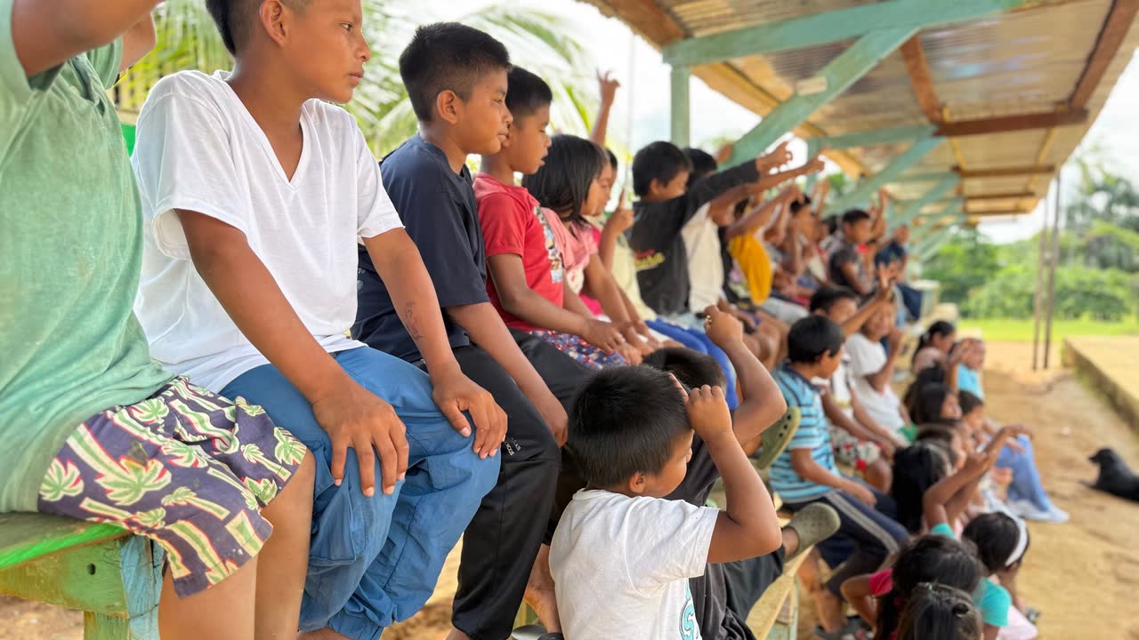 Niñas y niños sentados en una grada bajo el techo, atentos a la actividad