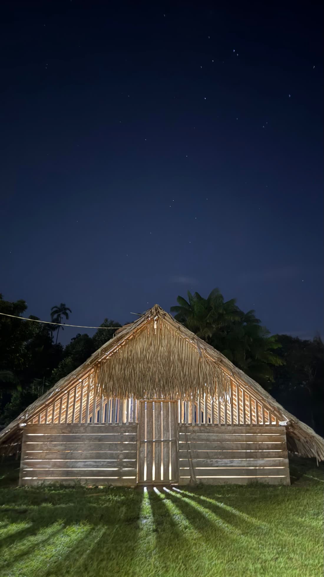 Maloca de techo de palma iluminada por dentro bajo un cielo estrellado